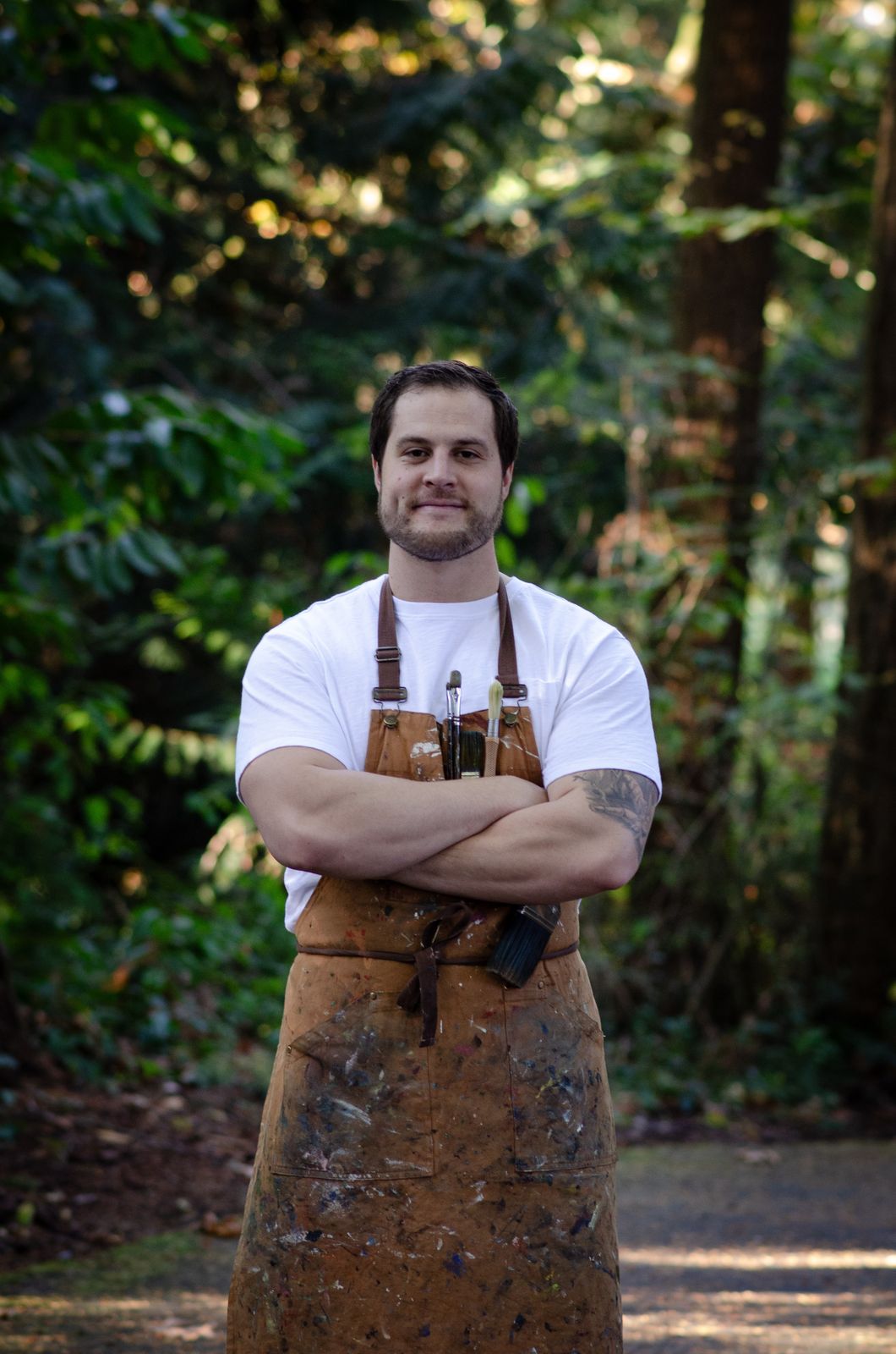 Gabe Johnson standing among evergreen trees, arms crossed, wearing a white shirt and a paint-covered canvas apron.