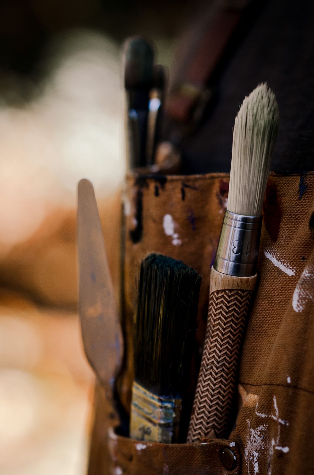 Brushes and a palette knife tucked into the pocket of a paint-spattered leather apron.